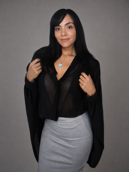 A woman with long black hair wearing a sheer black long-sleeve top over a black cardigan, paired with a light gray pencil skirt, standing against a solid gray studio background.